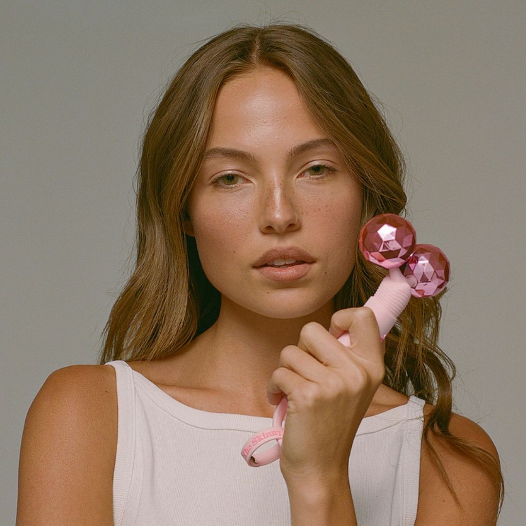 Woman in a white tank top holding a pink facial massage roller with two shiny pink spheres
